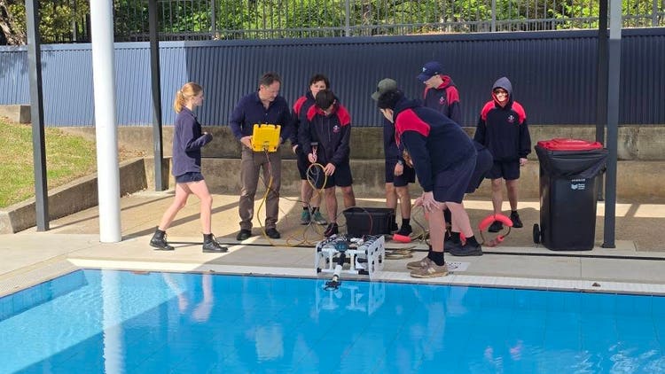 Seven students with teacher stand on side of swimming pool to launch their ROV submarine