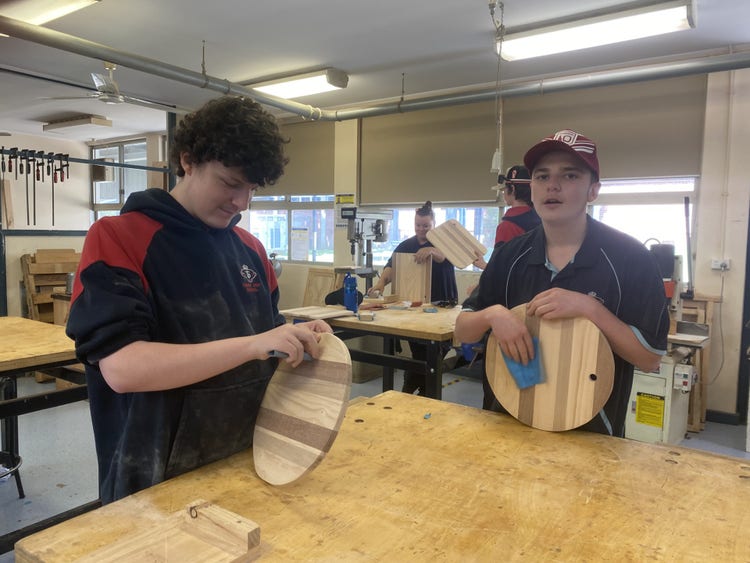 Two male students sand paper their chopping boards made in woodwork room with teacher in the background